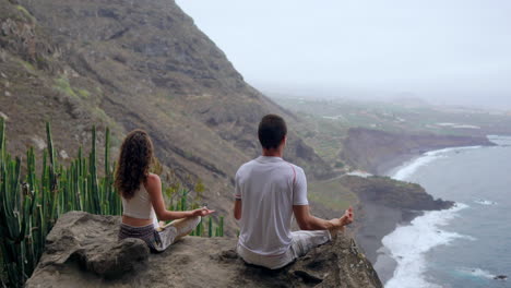 Positioned-atop-a-mountain,-a-man-and-woman-meditate-on-rocks,-raising-their-hands,-as-they-enjoy-the-ocean-panorama-and-engage-in-calming-breaths